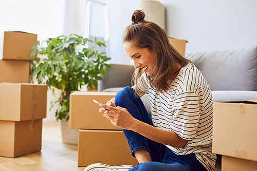 Beautiful young woman on smart phone while moving to new apartment. Surrounded by boxes